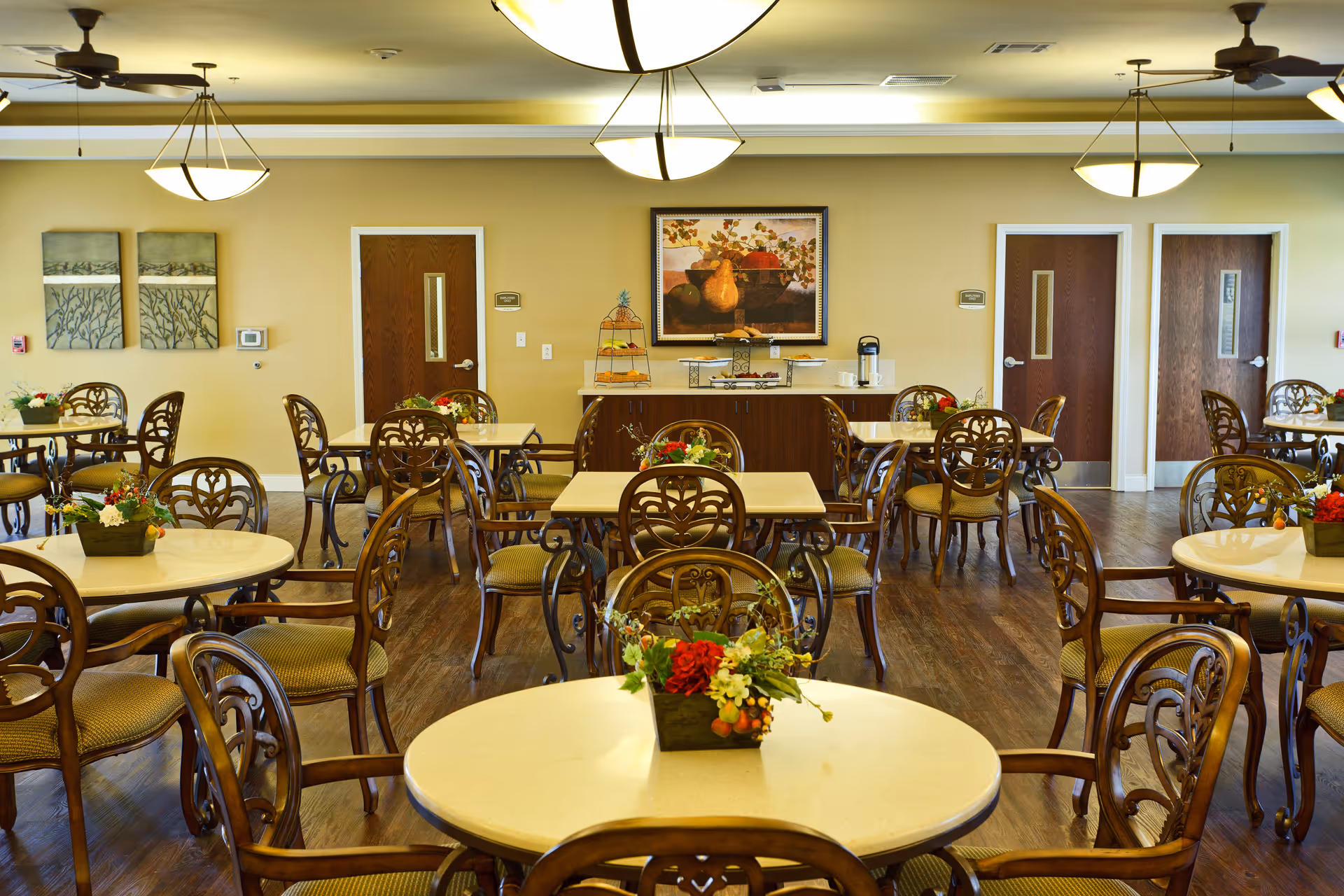 A well-lit dining room with multiple round and square tables surrounded by ornate wooden chairs with cushioned seats. Each table has a small floral centerpiece. The room has wooden flooring, ceiling fans, and modern hanging light fixtures. There are three wooden doors along the back wall, a sideboard with a coffee pot and tiered trays, and framed artwork on the walls.