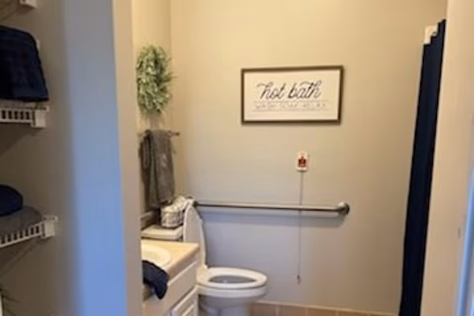 A bathroom with a white toilet, a sink with a countertop, and a towel rack with a gray towel. There is a framed sign on the wall above the toilet that reads 'hot bath' and a grab bar installed on the wall. Shelves with folded towels are visible on the left side.