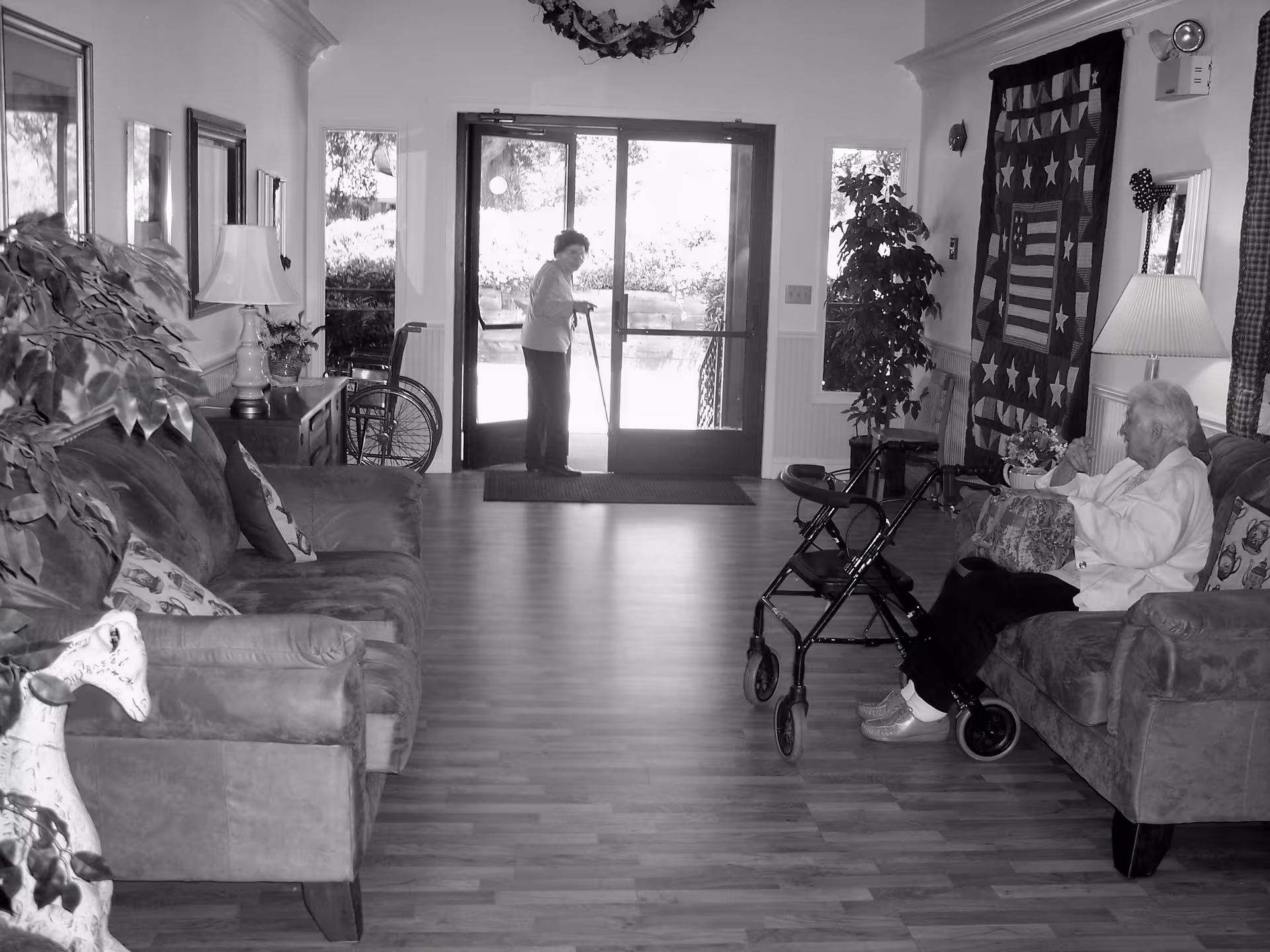 Black and white image of a retirement facility lounge area with two elderly women. One woman is standing near the glass exit doors using a cane, and the other is sitting on a couch with a walker in front of her. The room has sofas, a lamp, a plant, and a quilt with stars hanging on the wall.