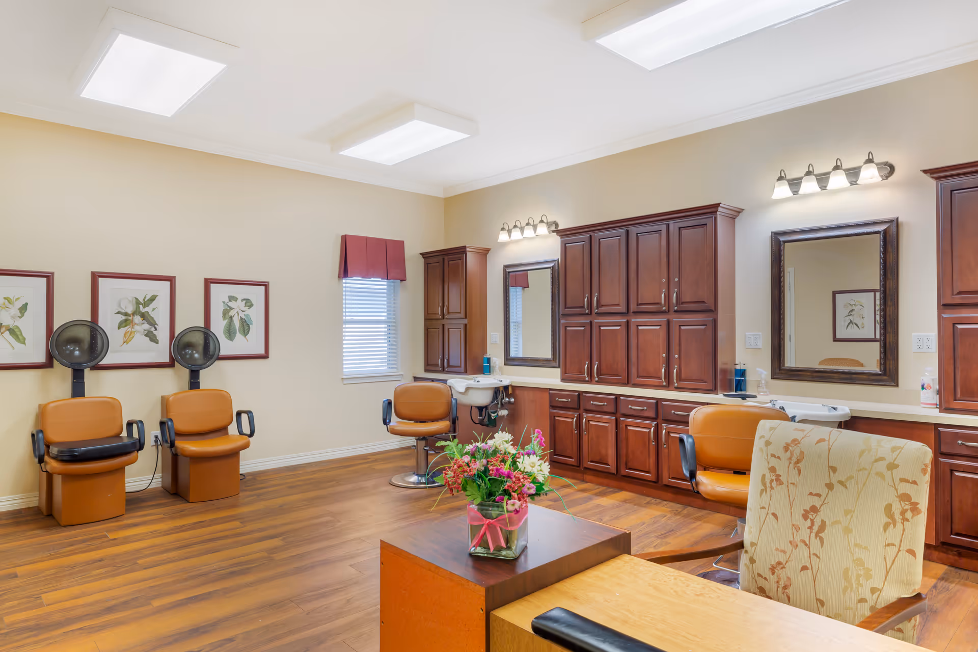 Interior view of a salon area in a senior living facility with wooden flooring, beige walls, and ceiling lights. The room features three orange salon chairs, two with hair dryers attached, and one in front of a sink. There are large wooden cabinets and two mirrors on the wall. A small table with a floral arrangement is in the foreground, along with a floral patterned armchair.
