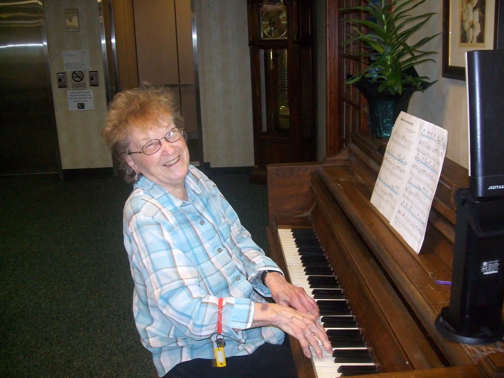 An elderly woman with glasses and a plaid shirt is sitting at a wooden piano, smiling while playing. There is sheet music on the piano and a potted plant on top. The background shows an elevator and a grandfather clock.