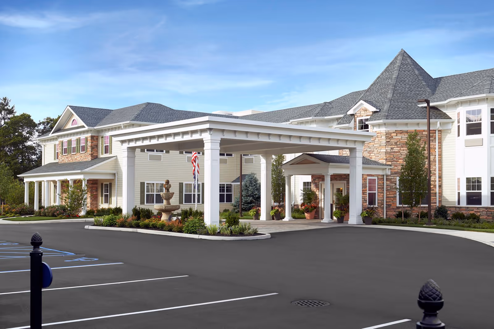 Exterior view of The Bristal Assisted Living at Sayville building featuring a covered entrance with white pillars, a stone fountain, landscaped greenery, and a clear blue sky.