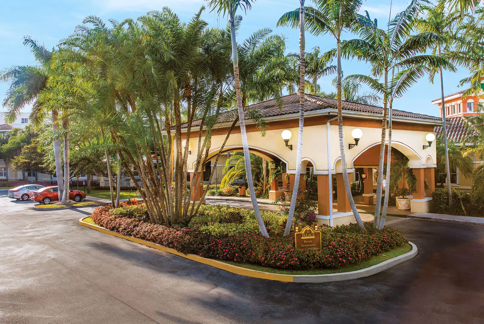 Porte-cochere entrance of a Mediterranean-style senior living building with palm trees, landscaped island, and parked cars.