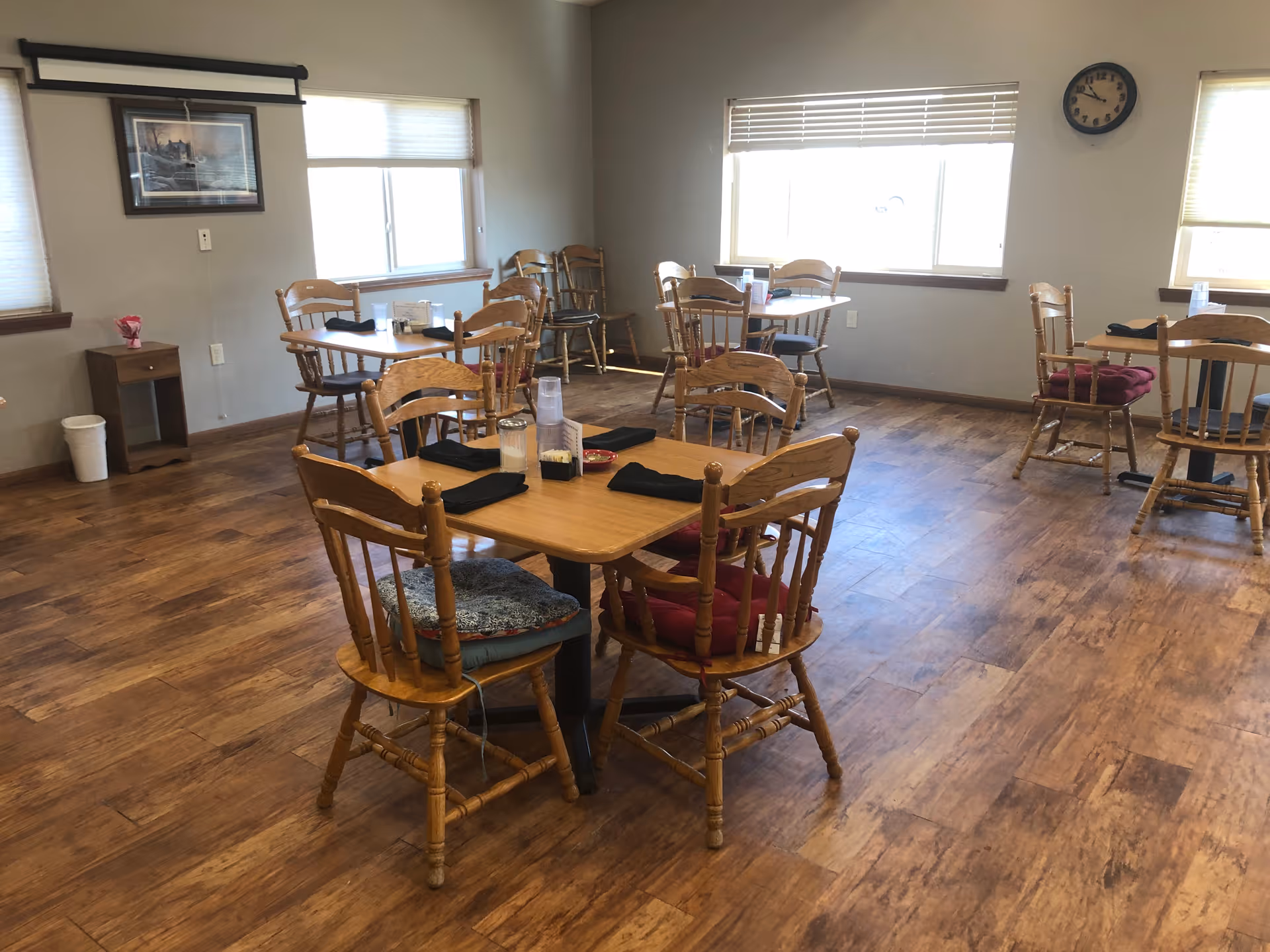 A dining room with wooden floors and several wooden tables and chairs. Each table has black napkins and some have cushions on the chairs. There are windows with blinds letting in natural light, a wall clock, and a framed picture on the wall.