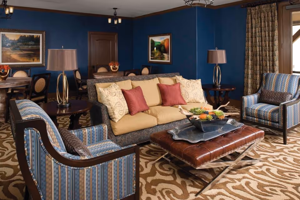 A cozy living room with a beige sofa adorned with red and cream pillows, two blue striped armchairs, a brown leather ottoman with a decorative tray and floral centerpiece, two side tables with lamps, blue walls with framed paintings, and a patterned carpet.