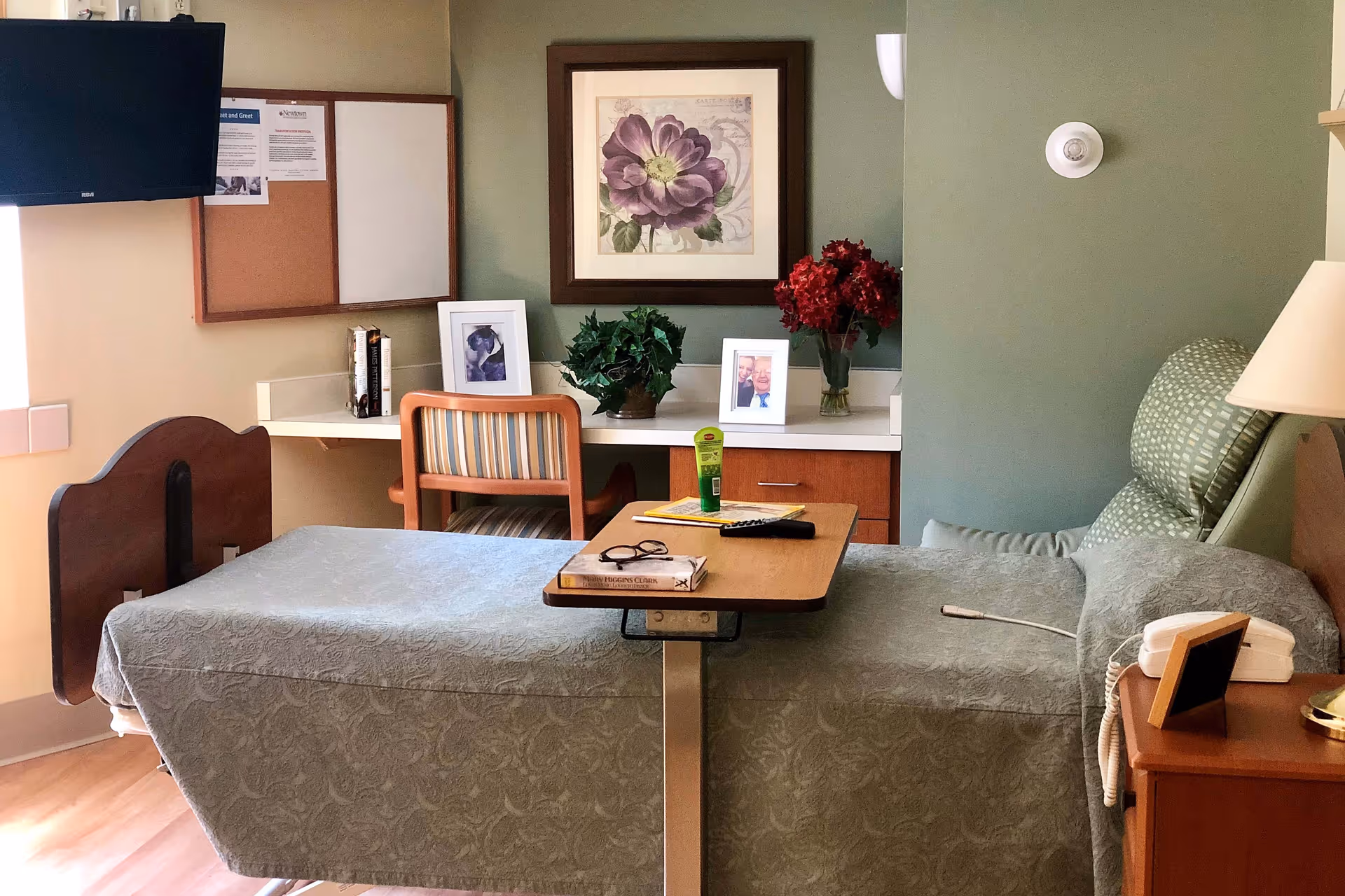 A tidy senior care patient room with a single bed, overbed table, desk and chair, TV, framed artwork and floral decorations.