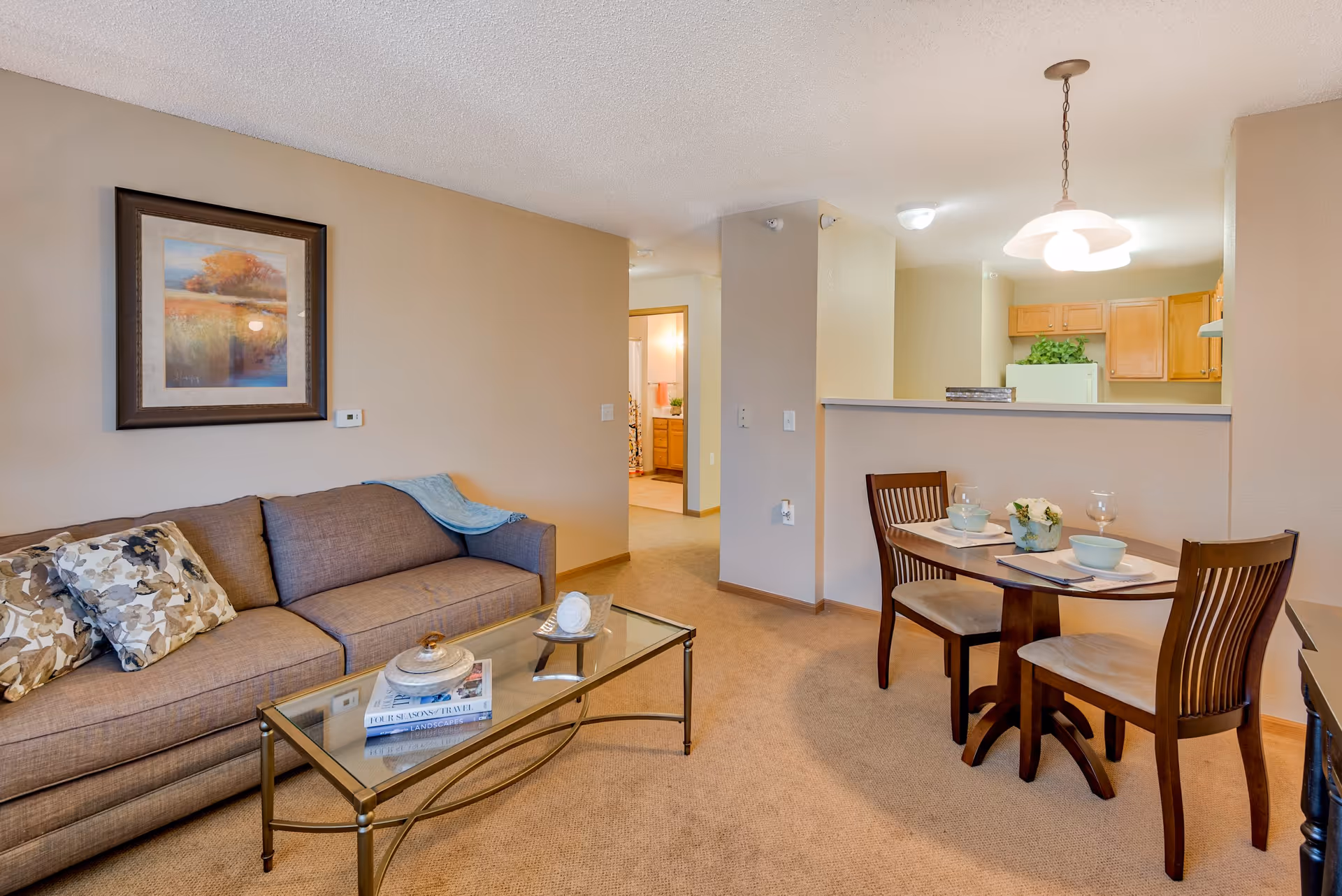 A cozy living and dining area in a senior living facility. The living room features a brown sofa with patterned cushions and a glass coffee table with decorative items. Adjacent to the living room is a small dining area with a wooden table set for two, including plates, bowls, and wine glasses. The kitchen is partially visible behind a half wall, with wooden cabinets and a refrigerator. The walls are painted beige, and a framed landscape painting hangs above the sofa. The carpet is light brown, and a doorway leads to another room, possibly a bathroom.
