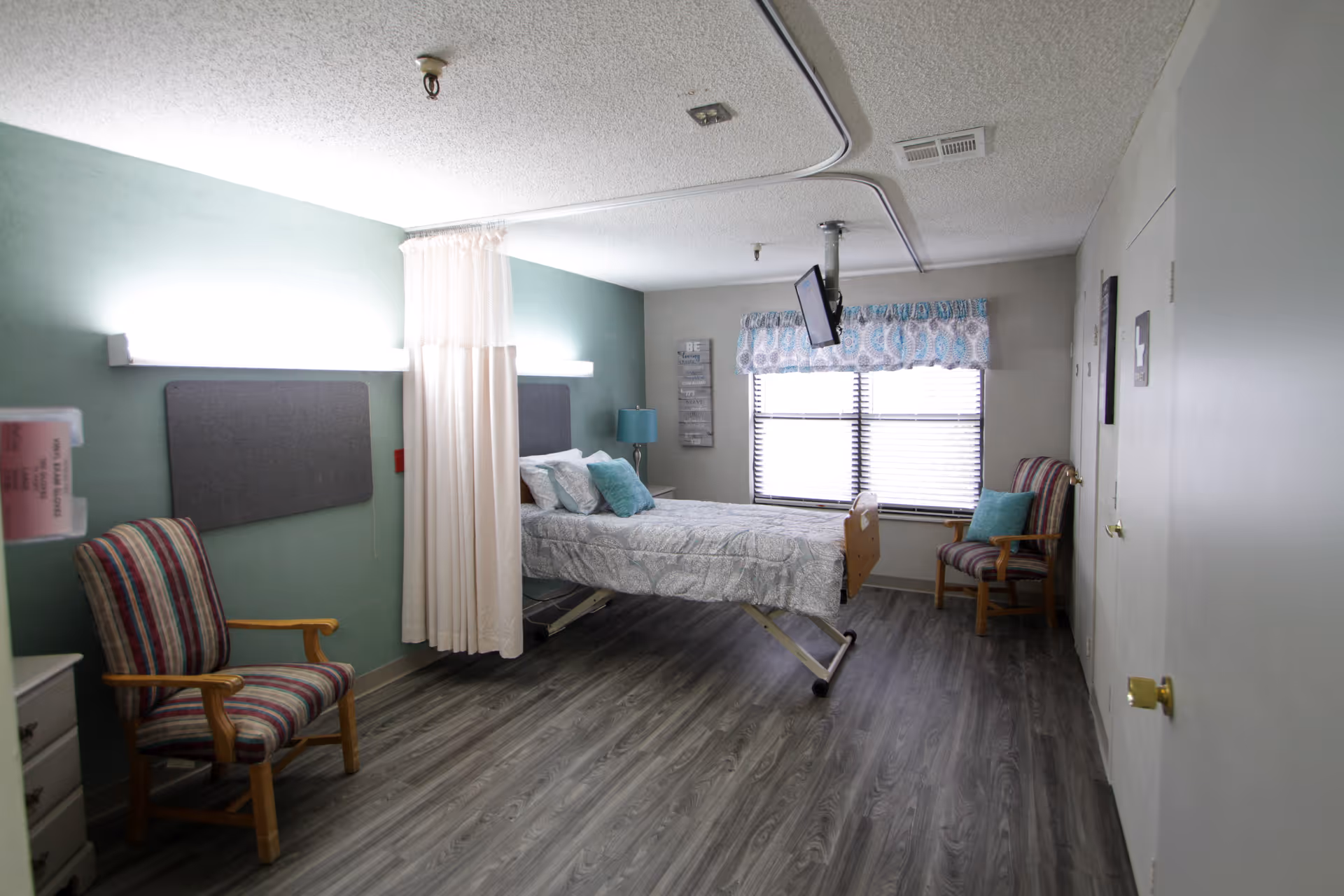 A hospital-style room with a single adjustable bed positioned near a window with blinds and a patterned valance. The room has two striped armchairs with wooden arms, one on each side of the bed. There is a curtain rail with a white privacy curtain partially drawn around the bed. The walls are painted light green and white, and the floor is covered with gray wood-patterned vinyl. A small TV is mounted from the ceiling near the window, and there is a blue lamp on a small table next to the bed.