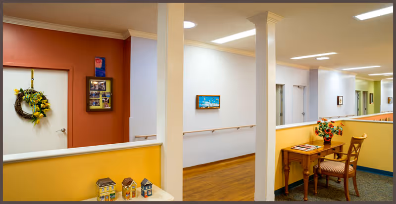 Interior hallway of a senior living facility with yellow and orange walls, wooden flooring, handrails along the walls, a small table with a vase of flowers and a chair, and decorative items including a wreath on a door and small model houses on a ledge.