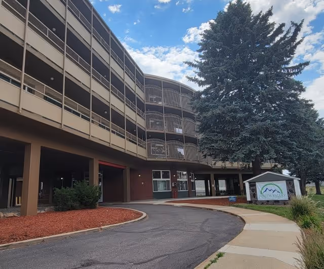 Curved multi-story rehabilitation facility facade with a covered driveway, large evergreen tree, and a sign reading 'South Valley' at the entrance.