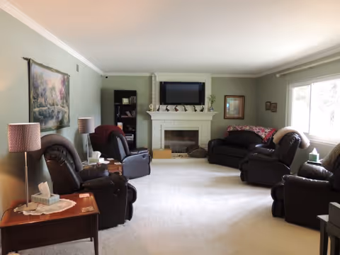 A cozy living room with light green walls and white carpet. The room features a white brick fireplace with a flat-screen TV mounted above it. There are four dark leather recliners and one dark sofa arranged around the fireplace. Two side tables with lamps and tissues are placed beside the recliners. A large window on the right side lets in natural light. A bookshelf and framed artwork decorate the walls.