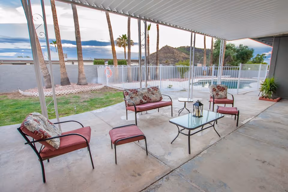 Covered outdoor patio area with metal framed chairs and a loveseat with red cushions and floral pillows, a glass coffee table with a lantern on top, and a small side table. Beyond the patio is a fenced swimming pool, palm trees, and a hill in the background under a cloudy sky.