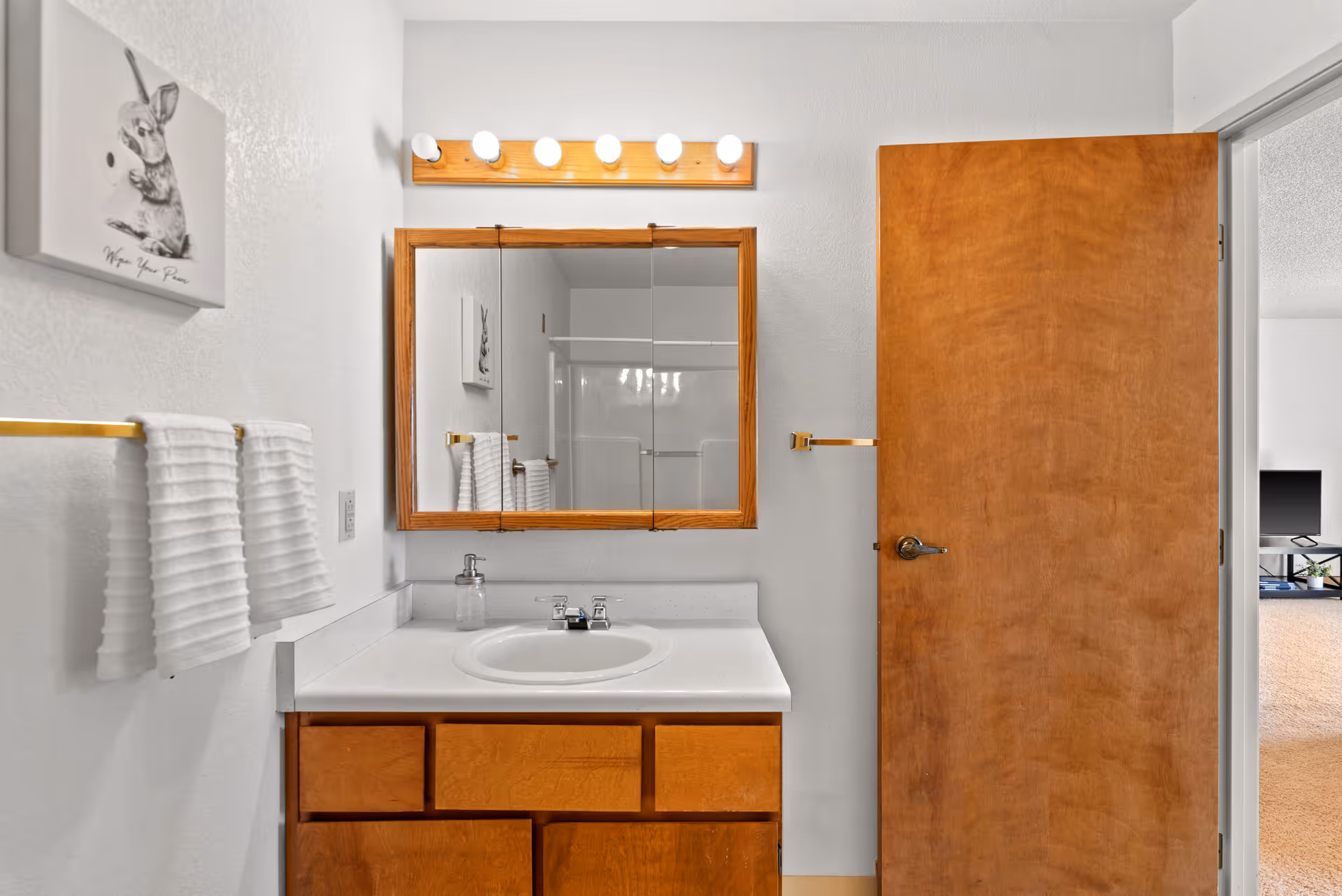 A bathroom vanity with a sink, wood-framed mirror and cabinets, light fixtures above, towel rack on the wall, and an open wooden door showing part of an adjacent room.