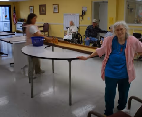 An indoor room with elderly people engaged in an activity involving a long wooden shuffleboard table. One elderly woman in a pink cardigan and blue shirt stands near the camera, smiling. Another person is walking near the table, and two elderly individuals are seated in the background, one in a wheelchair. The room has yellow walls with framed pictures and an American flag near the door.