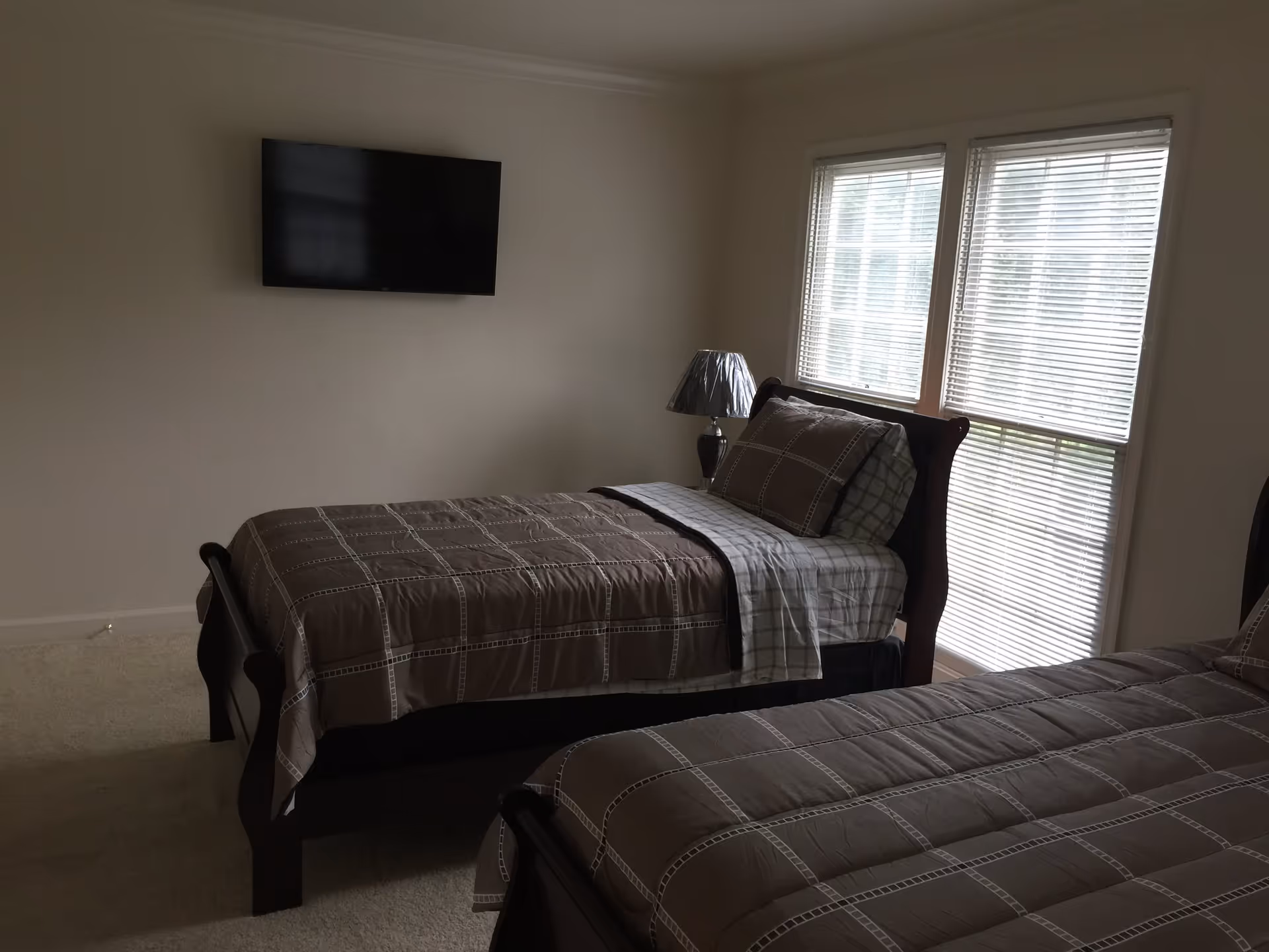 A bedroom with two single beds featuring matching brown and beige checkered bedding. There is a bedside table with a lamp between the beds, and a flat-screen TV mounted on the wall. Two large windows with blinds allow natural light into the room.