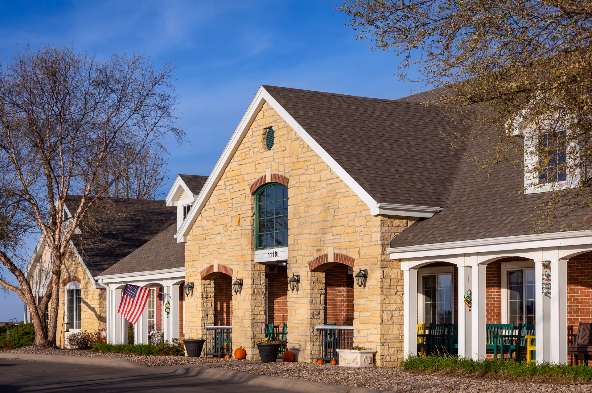 Exterior view of a single-story assisted living facility building with stone and brick facade, a pitched roof, and a covered porch with chairs. An American flag is displayed near the entrance, and there are some pumpkins and planters along the walkway. Trees with sparse leaves are visible around the building under a clear blue sky.