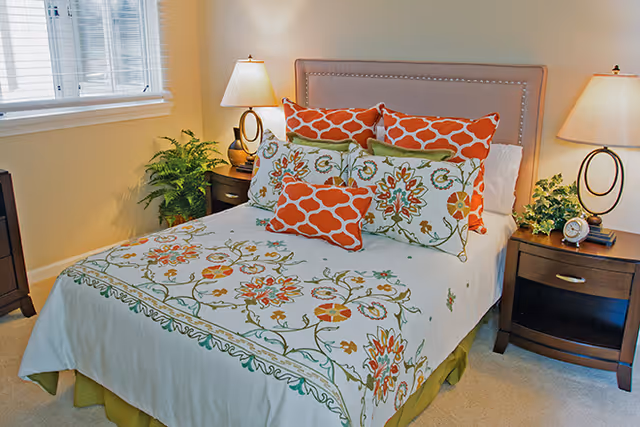 A neatly made bed with a floral and patterned bedspread and multiple decorative pillows in orange and green tones. The bed has a beige upholstered headboard with nailhead trim. There are two wooden nightstands on either side of the bed, each with a lamp and a small plant. A window with white blinds is visible on the left side of the room.