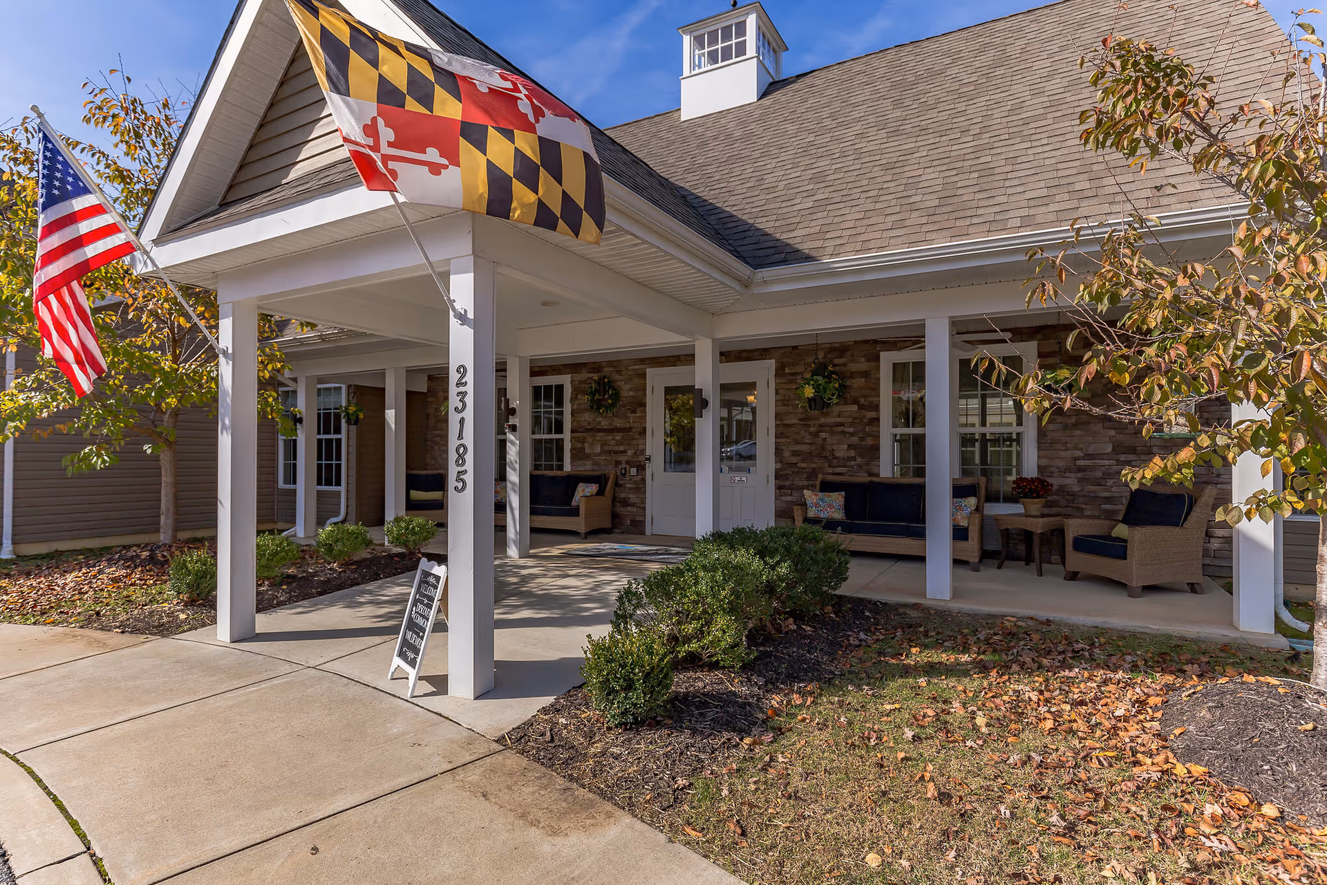 Entrance of a senior living facility with a covered porch featuring seating areas with cushioned chairs and sofas. Two flags, the American flag and the Maryland state flag, are displayed on poles attached to the porch. The building has a stone facade and white pillars, with the address number 23185 visible on one pillar. There are small bushes and trees with autumn leaves around the entrance.