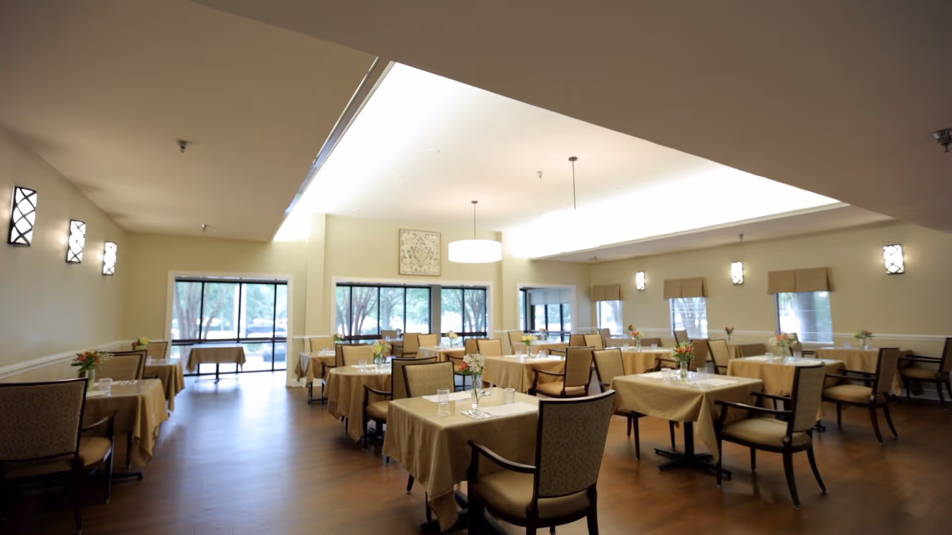 A spacious dining room with multiple tables covered in beige tablecloths, each set with glasses and small floral centerpieces. The room has large windows allowing natural light to enter, wall-mounted light fixtures, and a wooden floor.