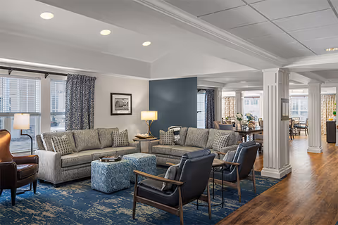 A spacious and well-lit living room area with two gray sofas, two dark armchairs, a patterned blue ottoman, and a blue patterned rug. The room features large windows with patterned curtains, a floor lamp, framed artwork on the wall, and wooden flooring extending into a dining area in the background.