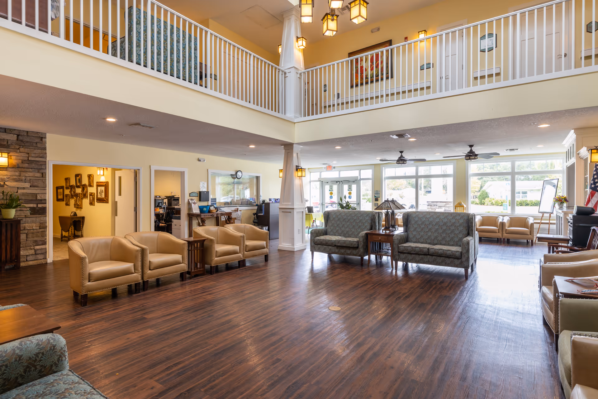 Spacious senior living facility common area with multiple seating arrangements including beige leather chairs and patterned sofas. The room features wood flooring, large windows allowing natural light, ceiling fans, and a second-floor balcony with white railings overlooking the area. There are decorative wall lights, framed artwork, and an American flag near the entrance.