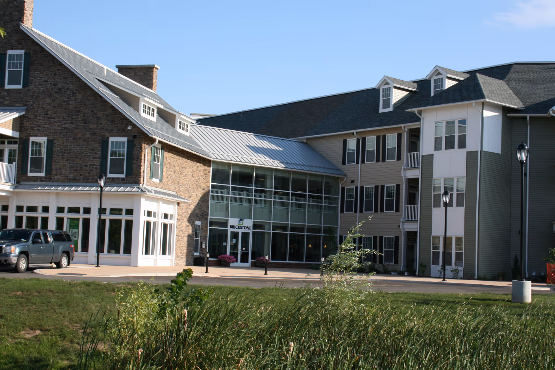 Front exterior of the Brickstone by St. John's senior living building with a glass entrance, three-story facade, parked SUV, and grassy foreground.