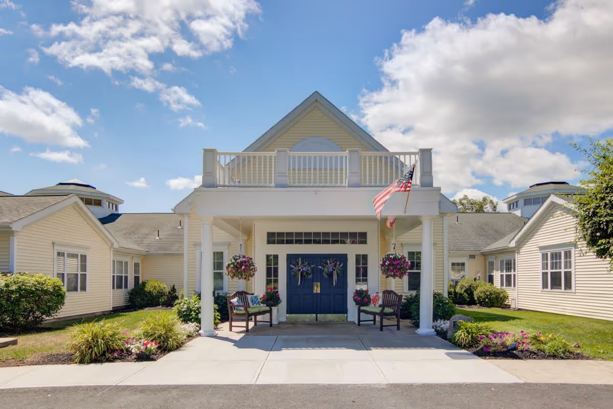 Front exterior view of The Atrium at Cardinal Drive building with a covered entrance, two benches, hanging flower baskets, an American flag, and well-maintained landscaping under a partly cloudy blue sky.