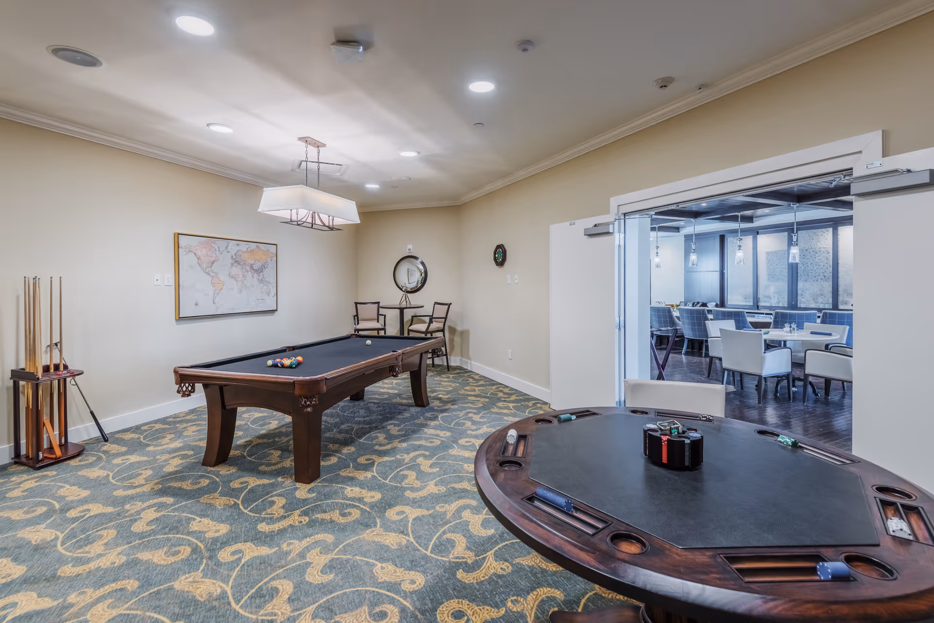 A recreational room featuring a pool table with billiard balls and cues, a poker table with poker chips and cards, two chairs with a small round table against the wall, a world map on the wall, and an adjacent dining area visible through an open doorway.