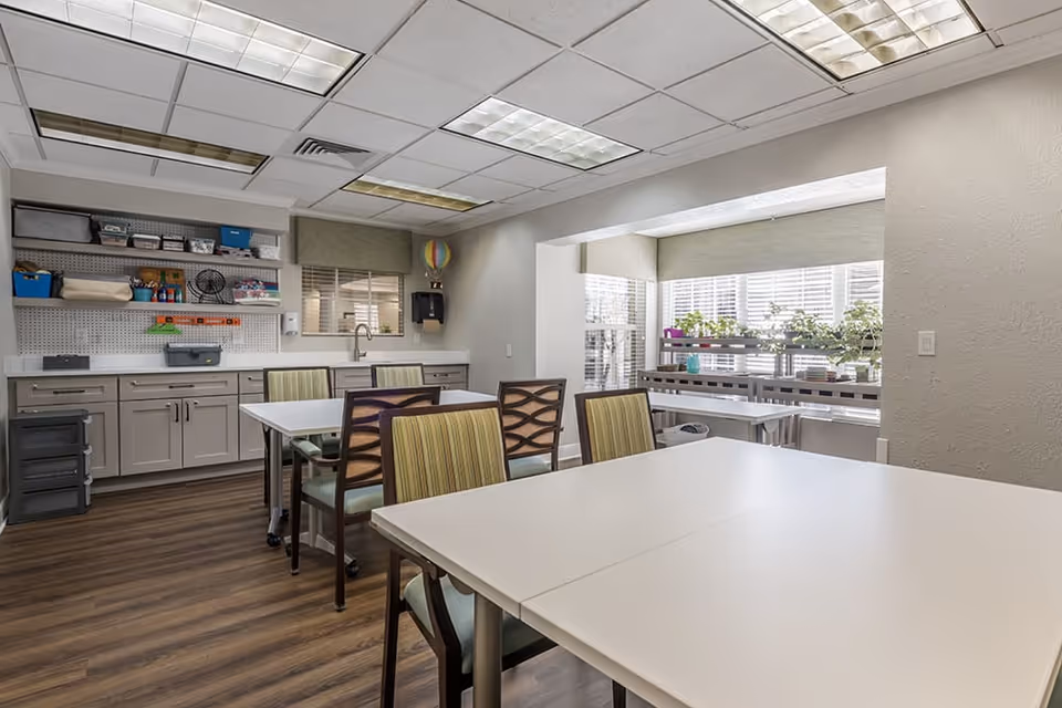 A well-lit room with white tables and chairs arranged for group activities or meetings. The room has a wooden floor, a countertop with cabinets, shelves holding various supplies, and a sink. A window with blinds and a small indoor garden with plants are visible in a recessed area.