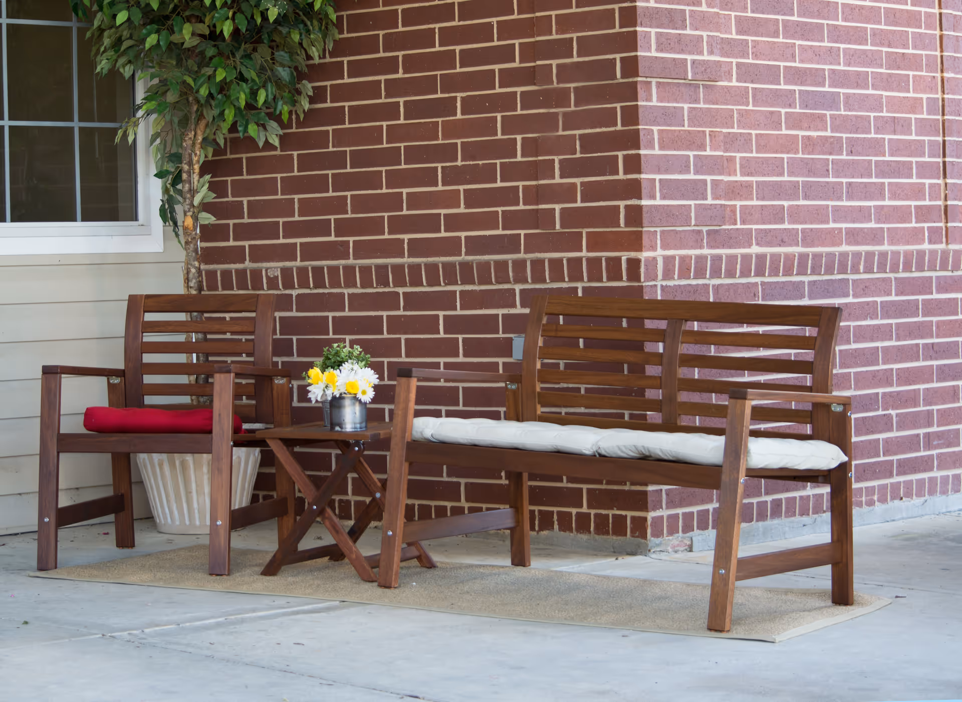 Outdoor seating area with wooden bench and chair, both with cushions, a small wooden table with a flower pot, a potted plant, and a brick wall background.