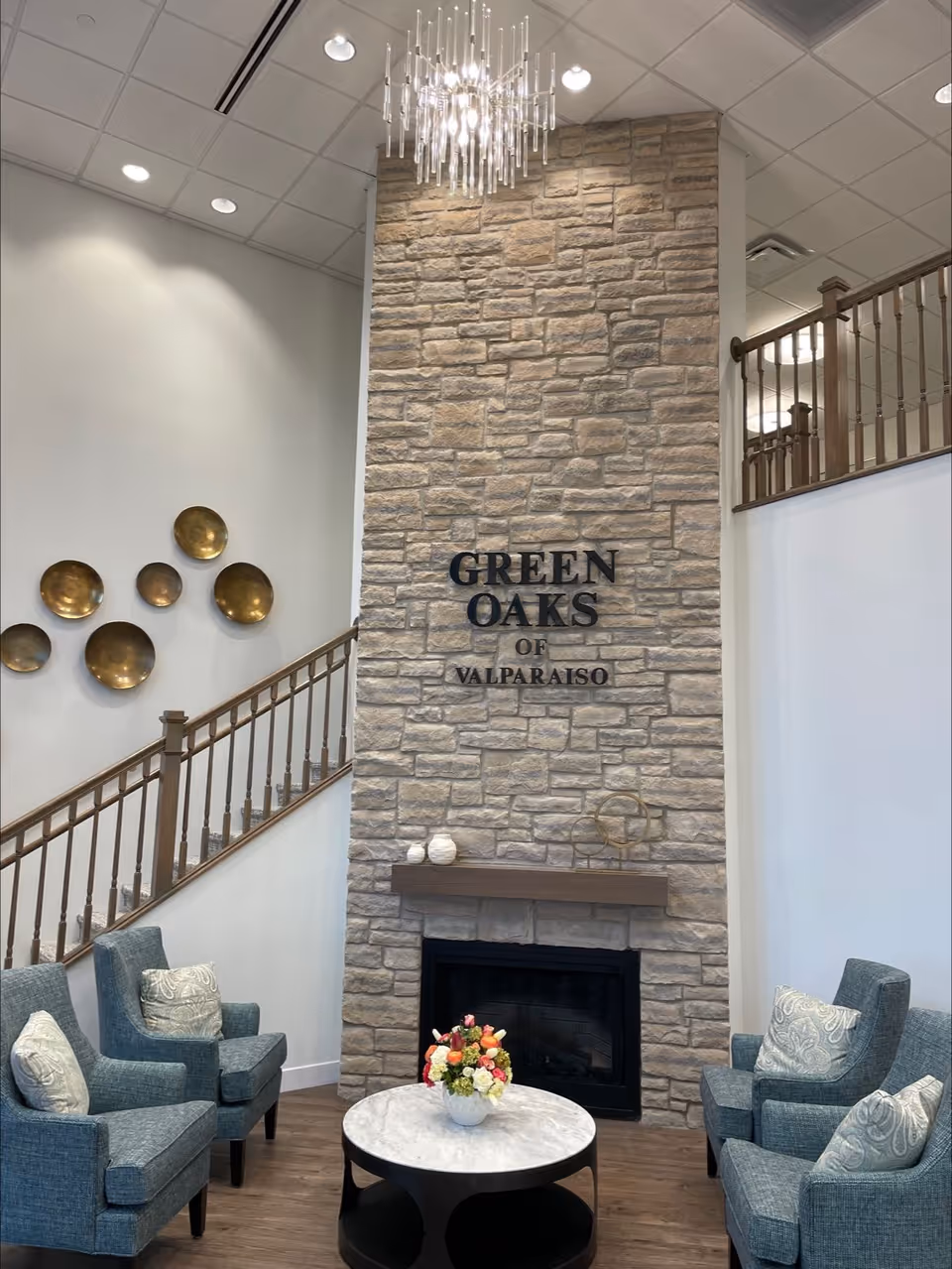 A cozy sitting area inside Green Oaks of Valparaiso featuring a tall stone fireplace with the facility name mounted on it. Four blue upholstered armchairs with patterned cushions are arranged around a round marble-top coffee table with a floral centerpiece. A modern chandelier hangs from the ceiling, and a wooden staircase with a railing is visible on the left side. Decorative gold wall plates adorn the wall above the staircase.