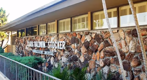 Stone-faced exterior wall of the Clara Baldwin-Stocker Home with the facility name mounted below a row of windows and a railing in front.