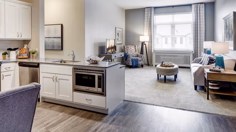 A modern open-concept living space featuring a kitchen area with white cabinets, a built-in microwave, and a sink island. Adjacent to the kitchen is a living room with a large window, a patterned armchair, a sofa with cushions, a round ottoman coffee table, and two side tables with lamps. The room is decorated with framed artwork and has a combination of wood flooring and carpet.