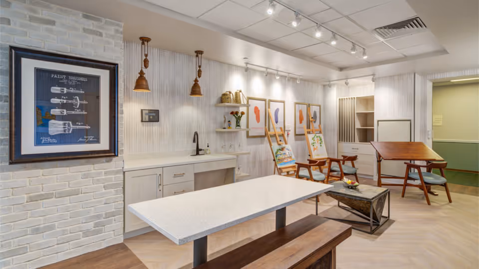 A bright and modern art studio room with a white countertop island and wooden bench in the center. The back wall features a sink, shelves with decorative items, and three easels holding paintings. There are three framed abstract art prints on the wall, pendant lights hanging from the ceiling, and a drafting table with a chair on the right side. The room has light-colored wood flooring and white walls with subtle vertical texture.