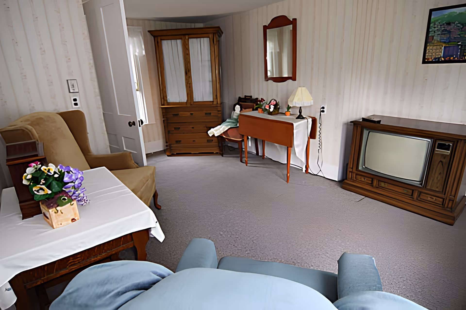 Cozy living room with a vintage TV console, armchairs, side tables with flowers, and a wooden cabinet against wallpapered walls.