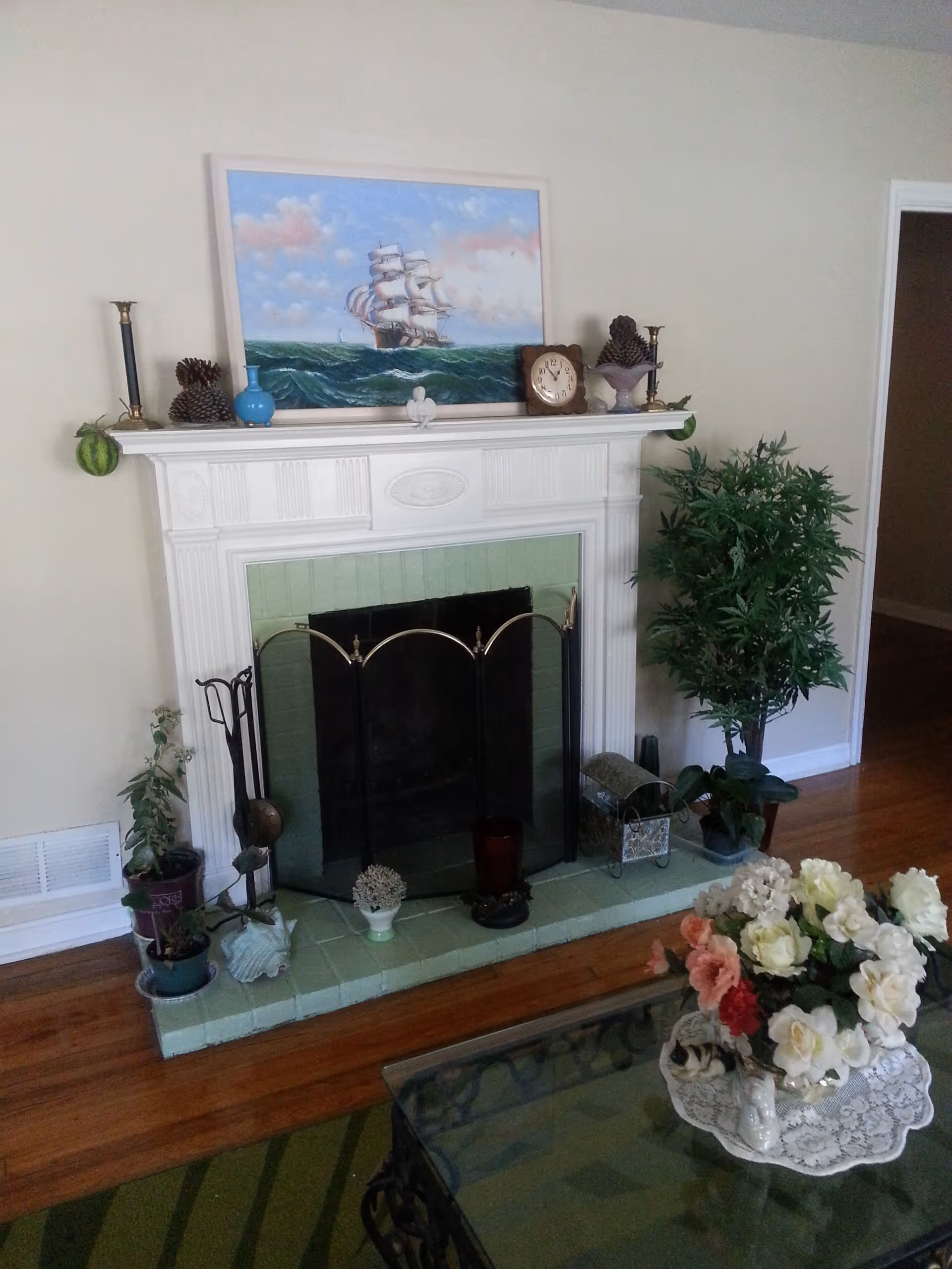A living room fireplace with a white mantel and green tile hearth, decorated with plants, ornaments and a ship painting above, and a coffee table with a floral arrangement in front.