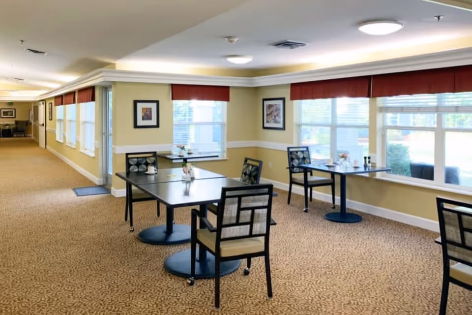 A bright dining area in a senior living facility with several square tables and cushioned chairs. The room has large windows with red valances allowing natural light to fill the space. The walls are painted light yellow with white trim, and framed artwork hangs on the walls. The floor is carpeted with a patterned design.