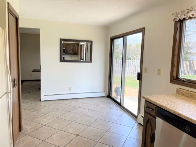 A bright kitchen area with tiled floor, wooden cabinets, a dishwasher, and a sliding glass door leading to an outdoor fenced yard. There is a window with a floral valance and a mirror on the wall reflecting part of the kitchen.