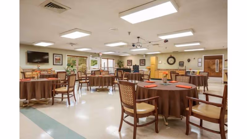 A spacious dining room with multiple round tables covered with brown tablecloths, each set with orange napkins and a small flower centerpiece. The room has beige walls, a tiled floor, ceiling lights, a wall-mounted TV, framed pictures, and a large clock on the far wall. Several wooden chairs with cushioned seats and backs are arranged around the tables.
