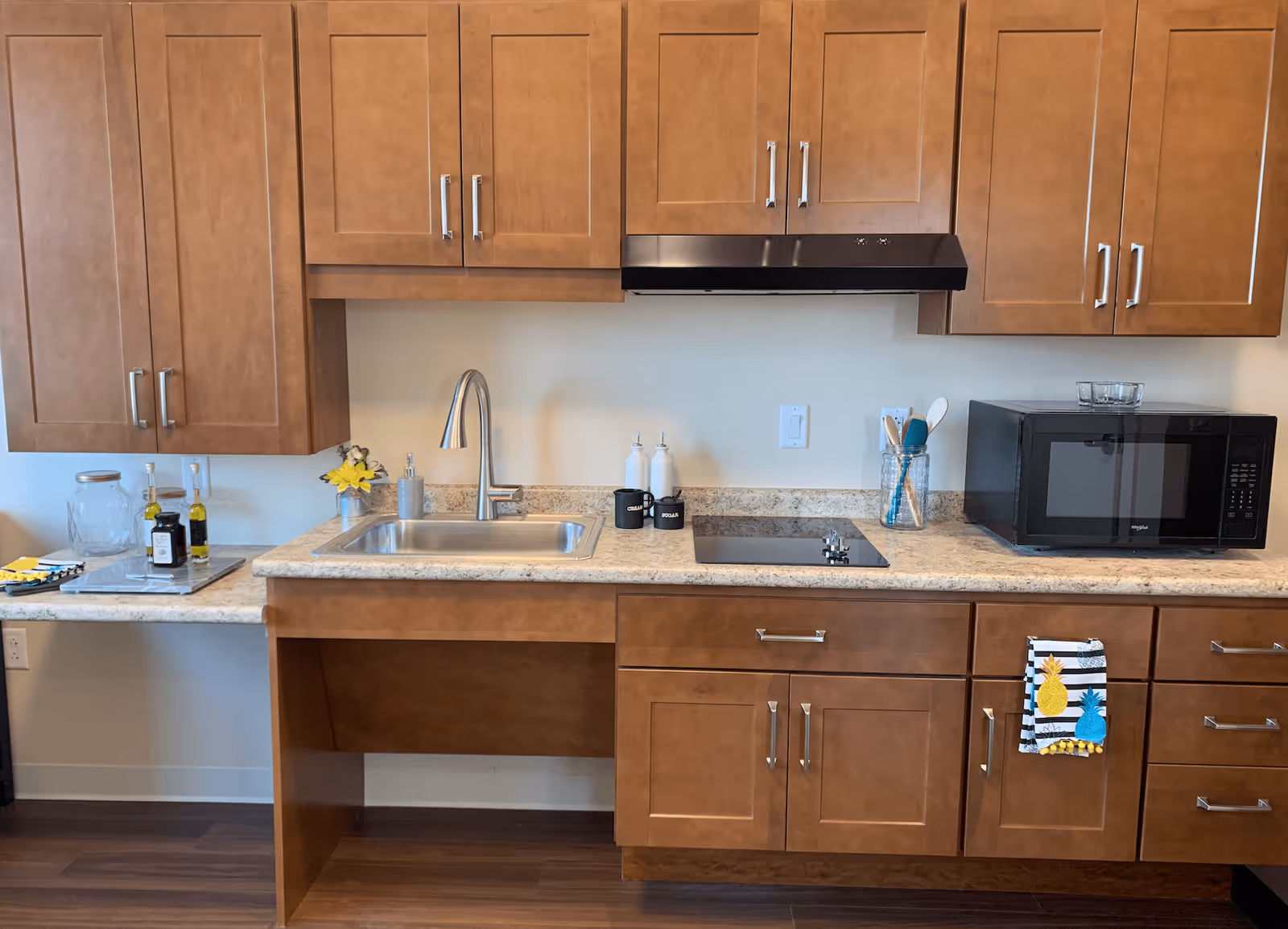 Kitchenette with brown wood cabinets, a stainless steel sink and faucet, an electric cooktop, microwave, and countertop accessories.