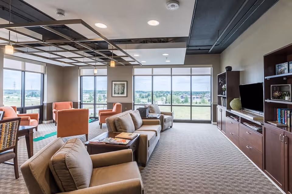 Bright lounge with sofas and armchairs, large windows overlooking a grassy landscape, and a TV on a wooden media wall.