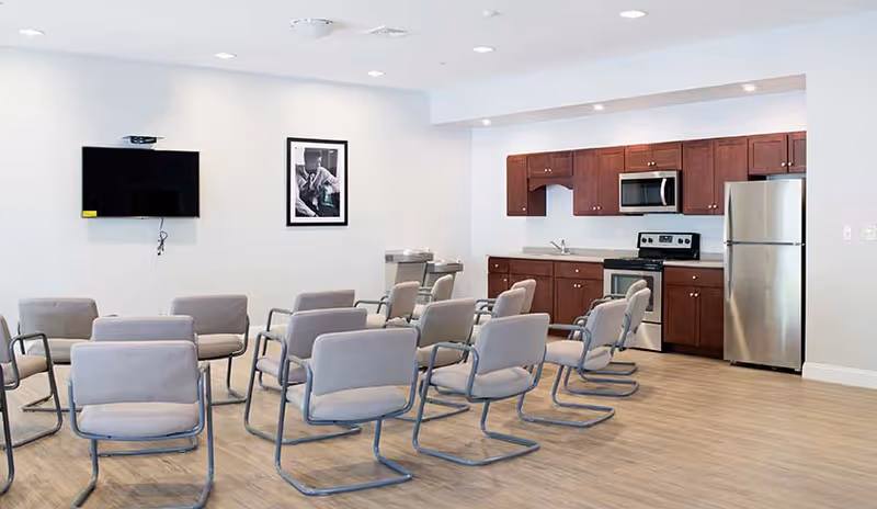 A room with multiple gray cushioned chairs arranged in rows facing a wall-mounted flat screen TV and a black and white framed photograph. The room also features a kitchenette area with wooden cabinets, a stainless steel refrigerator, microwave, stove, and sink. The floor is light wood, and the walls are white with recessed lighting in the ceiling.