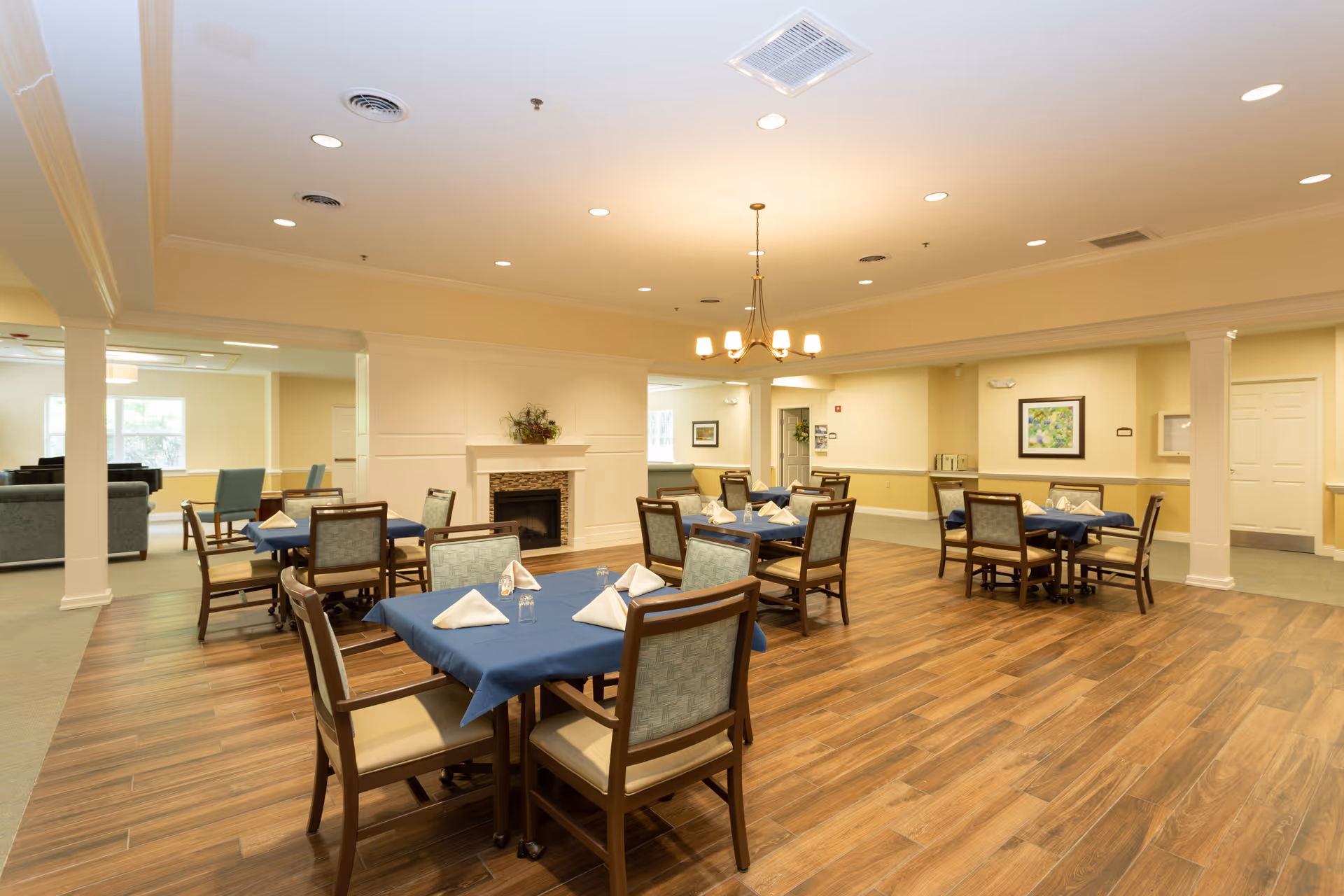 A spacious dining room with several tables covered in blue tablecloths, each set with folded white napkins and glasses. The room features wooden flooring, a chandelier hanging from the ceiling, a fireplace with a decorative plant on the mantel, and light yellow walls. There are chairs around each table and a seating area with sofas visible in the background.