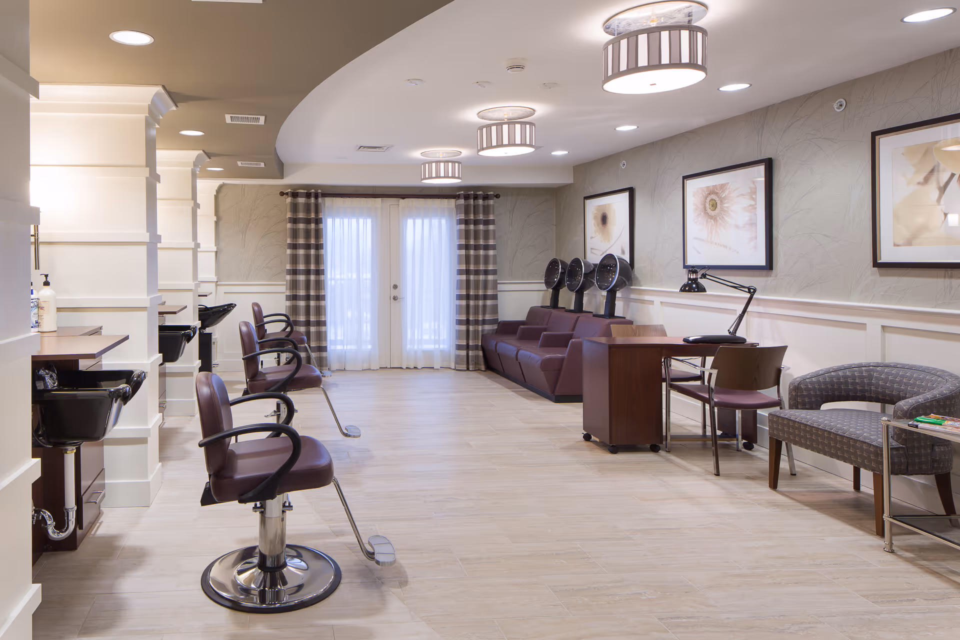 Interior view of a senior living facility's hair salon area with multiple salon chairs and hair dryers. The room has light-colored walls with framed floral artwork, a small desk with a lamp, and a seating area with a cushioned bench and a small table. The floor is light wood, and there are large windows with sheer curtains and plaid drapes at the far end.