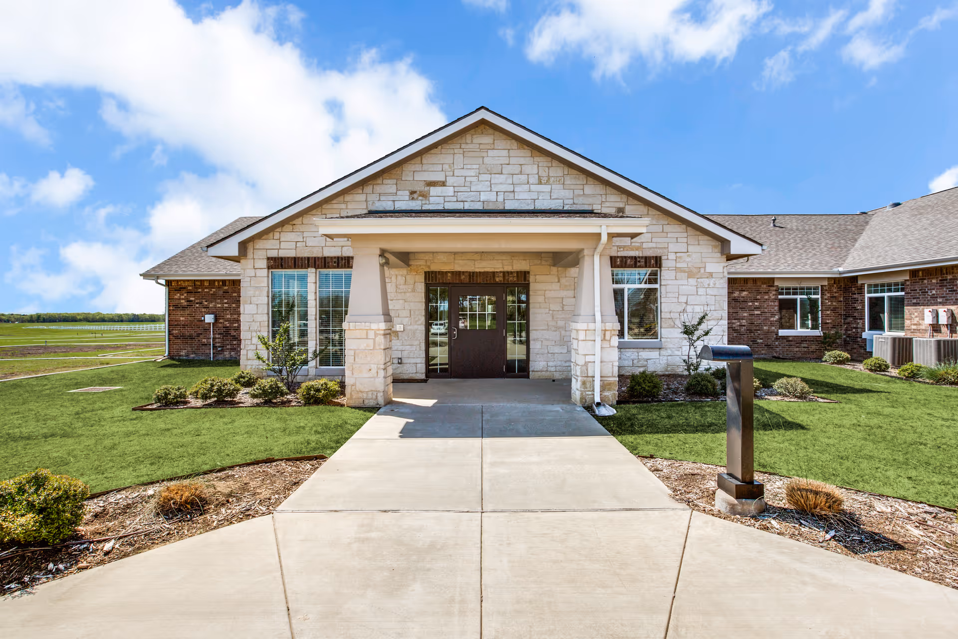 Front entrance of a single-story brick-and-stone building with a covered entry and concrete walkway through a landscaped lawn.