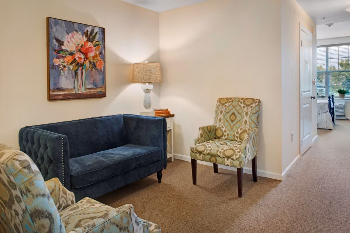 A cozy sitting area with a dark blue velvet sofa and two patterned armchairs on a carpeted floor. A small white side table with a lamp and some books is placed in the corner. A colorful floral painting hangs on the beige wall above the sofa. In the background, there is a hallway leading to a bedroom with a window letting in natural light.