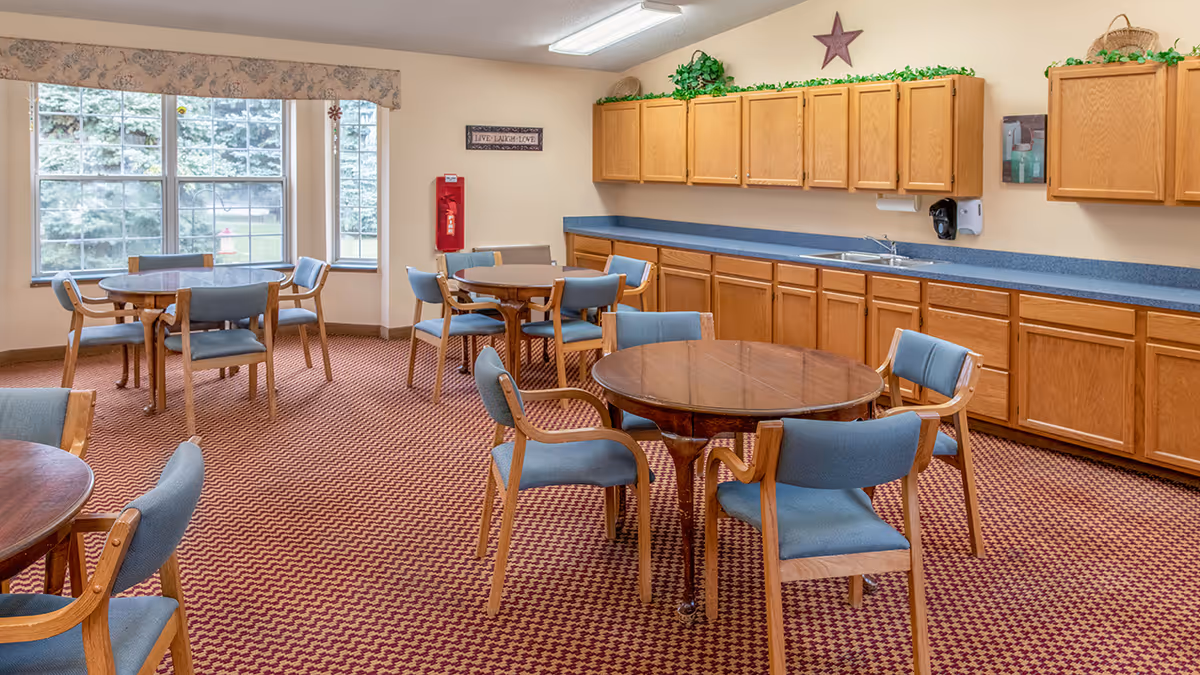 A dining room with round wooden tables and blue cushioned chairs arranged on a patterned carpet. The room has large windows with a view of greenery outside. Along one wall, there are wooden cabinets with a blue countertop and a sink. Decorative plants and a star adorn the top of the cabinets, and a sign on the wall reads 'LIVE LAUGH LOVE'.