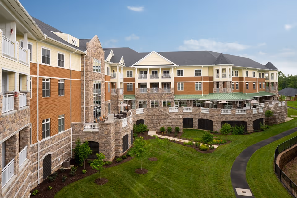 Exterior view of a large senior living facility building with a combination of stone and brick facade, multiple windows, balconies, and a green lawn with landscaped bushes and a paved walkway in front.