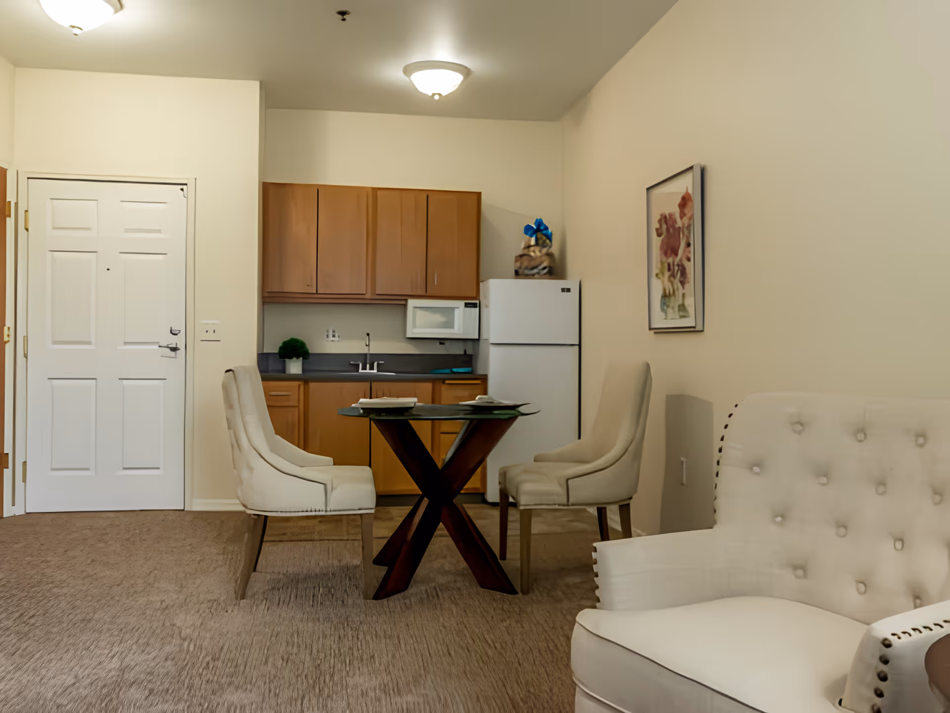 Interior view of a senior living facility room featuring a small kitchen area with wooden cabinets, a white refrigerator, and a microwave. In front of the kitchen is a glass-top dining table with two upholstered chairs. To the right, there is a white tufted armchair and a framed floral artwork on the wall. The room has beige walls and carpeted flooring.