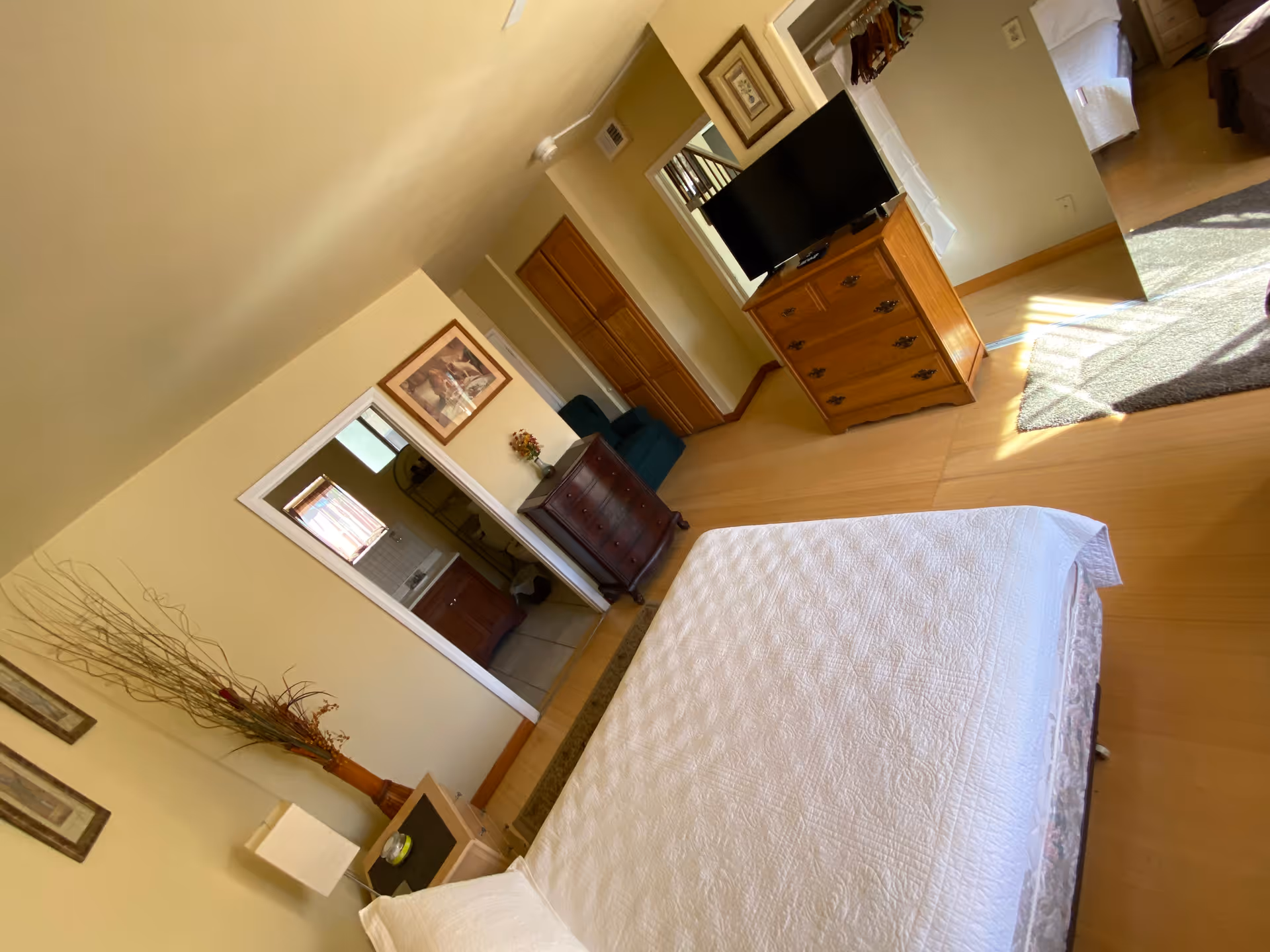 A bright bedroom with a large bed covered in a white quilt. There is a wooden dresser with a flat-screen TV on top, a small dark wood chest of drawers, and a green armchair in the corner. A doorway leads to a bathroom with a sink and mirror visible. The room has light-colored walls and wooden flooring, with sunlight streaming in from a window.